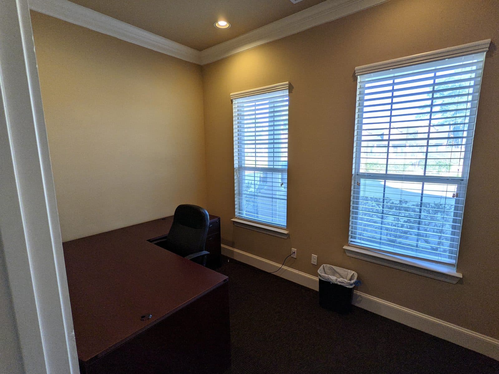 Compact private office with an L-shaped desk, a black task chair, a small file cabinet and two blinded windows looking out onto trees.
