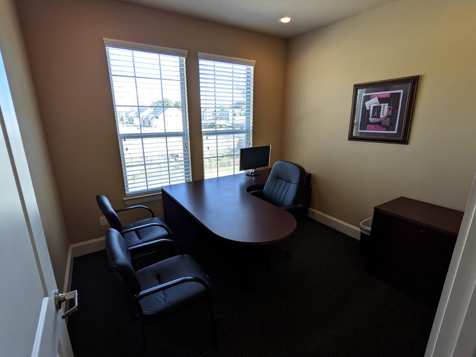 Private office with an L-shaped desk holding a flat-screen monitor, a black executive chair, two guest chairs, a wood filing cabinet, framed wall art, and two windows looking out onto houses and lawn.