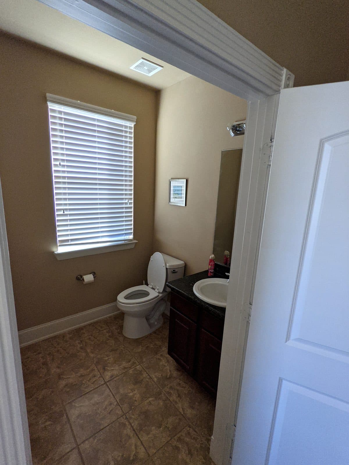 Private bathroom with a toilet, a dark wood vanity with a stone-look countertop and sink, a wall mirror, a blinded window and tiled flooring.