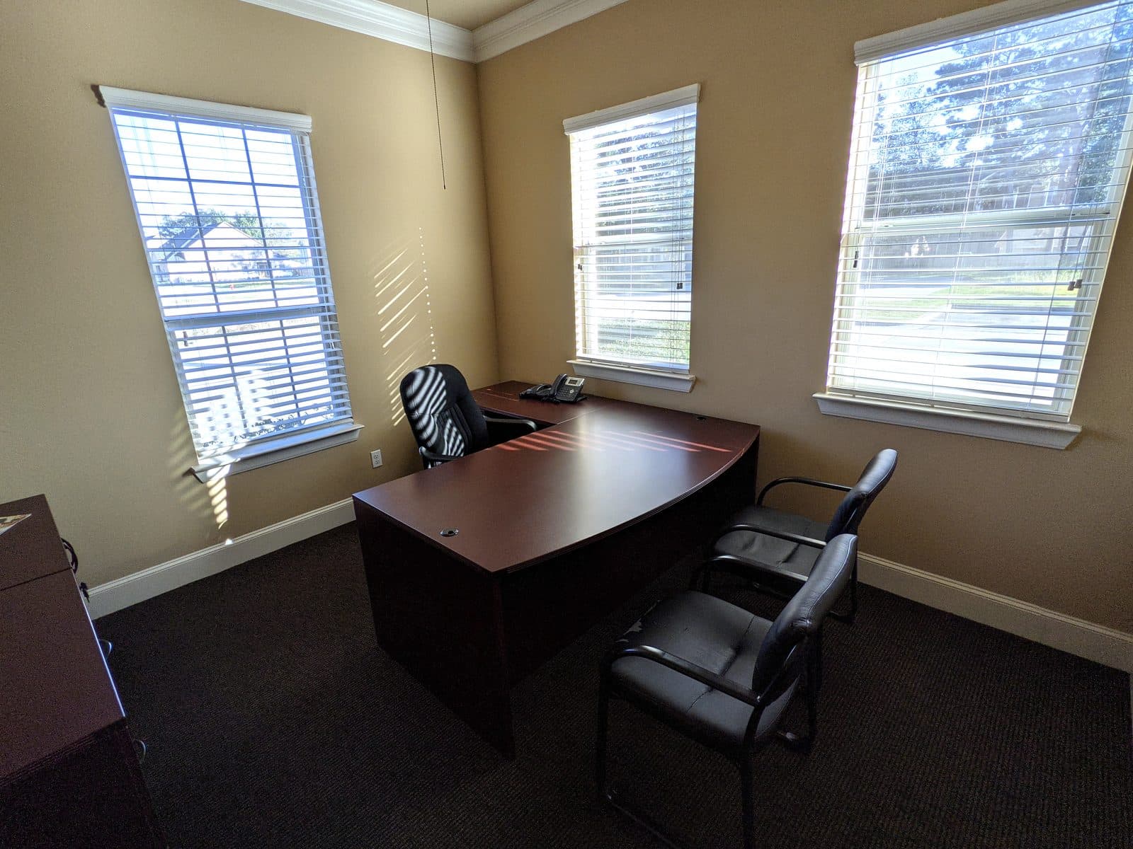 Corner of a private office with three windows, a bow-front mahogany desk and return, a desk phone, a black mesh task chair and two black guest chairs on dark grey carpet.