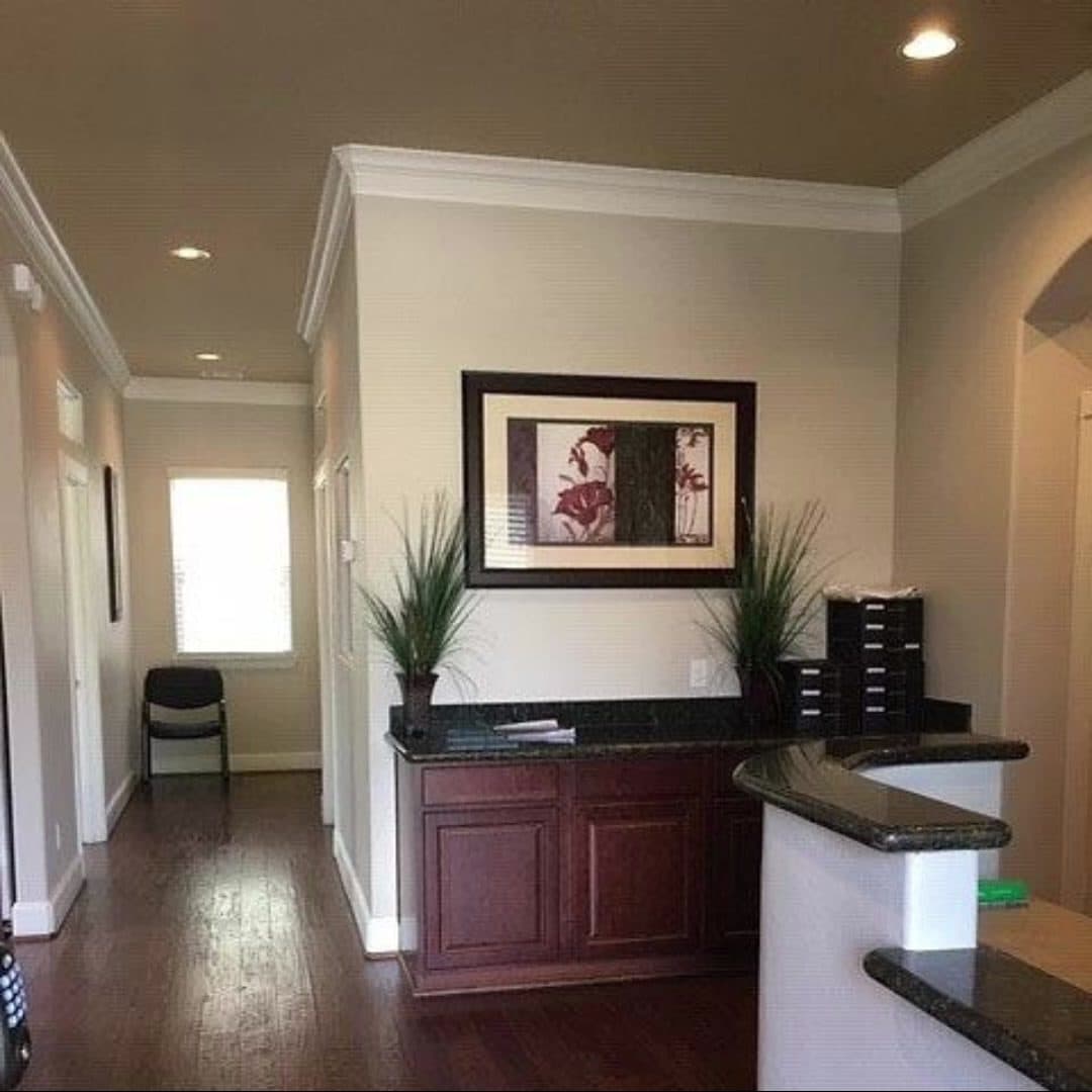 Reception area with a granite-topped counter over cherry cabinetry, framed floral artwork, artificial plants, hardwood flooring and a hallway leading to a window.